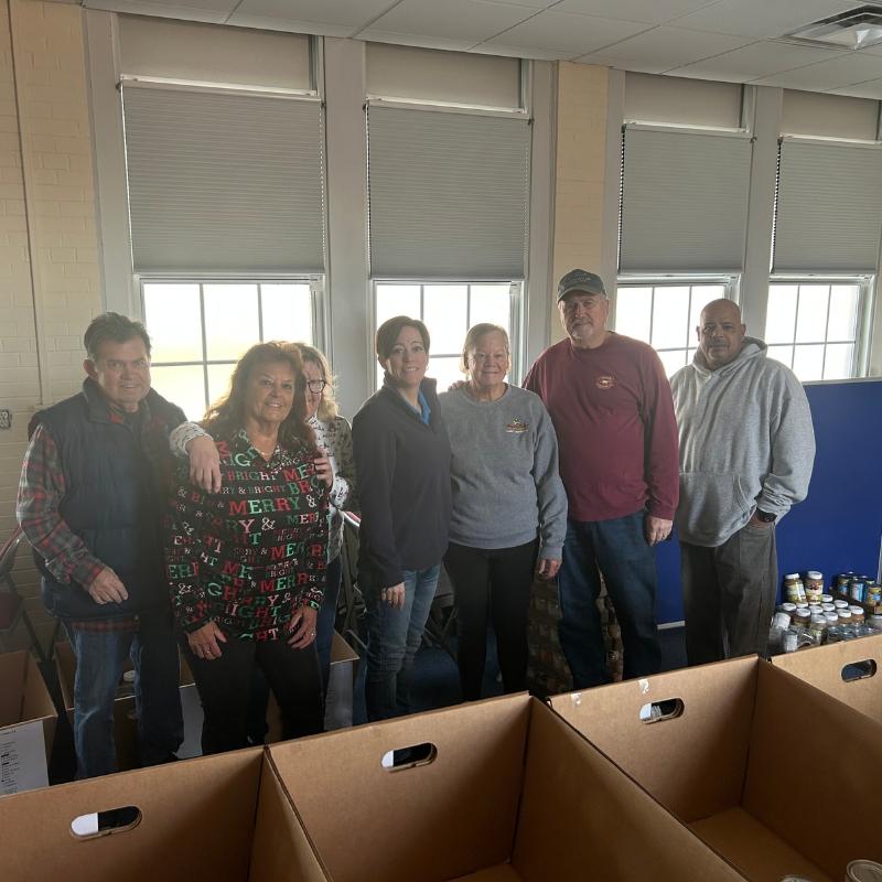 FD Food Closet 2 Group of seven volunteers posing in a room with large windows, standing behind empty brown donation boxes and a row of canned goods on the right side.