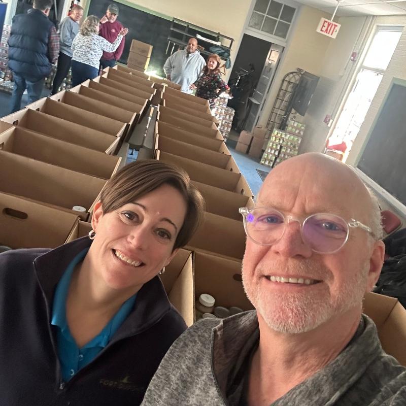 FD Food Closet3 Two people take a selfie in a room filled with long rows of open cardboard boxes while others work in the background at a packing drive.